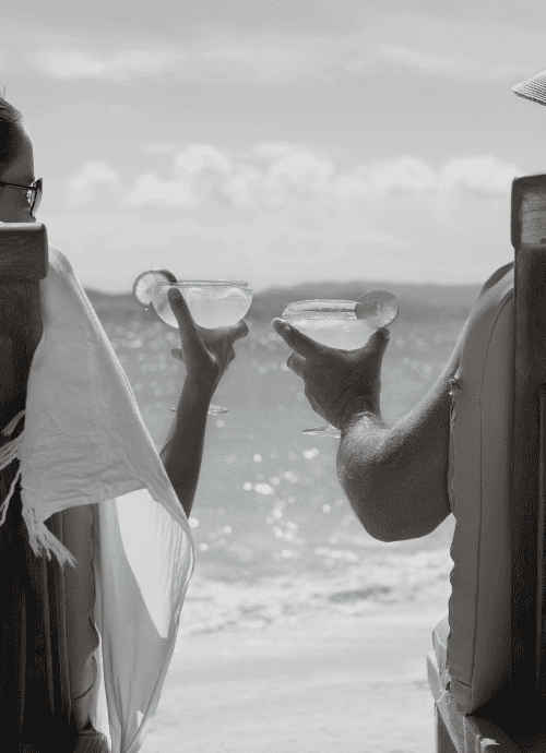 Black and white photo of a couple doing a cheers with cocktails sat on loungers on the beach